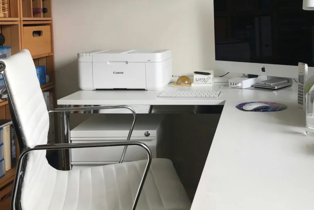 A white desktop printer beside a monitor and keyboard in an everyday home workspace.