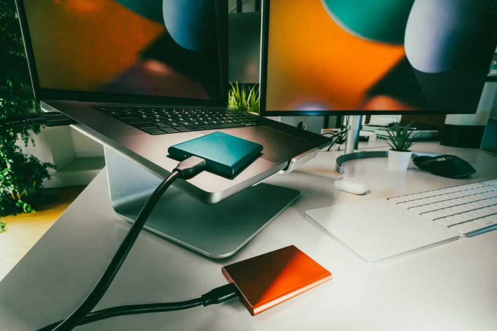 Two portable drives connected by cables beside a laptop on a stand and an external monitor.